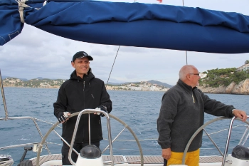 Zwei männliche Segler stehen am Steuerrad, im Hintergrund bergige Inselwelt