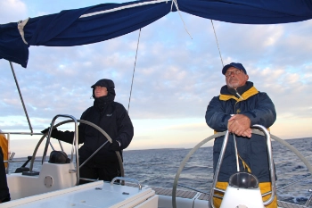 Ein Mann und eine Frau stehen am Steuerrad, im Hintergrund blaues Wasser